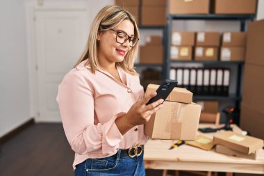 Young hispanic woman ecommerce business worker holding packages and using smartphone at office