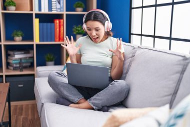 Hispanic young woman using laptop at home wearing headphones celebrating victory with happy smile and winner expression with raised hands 