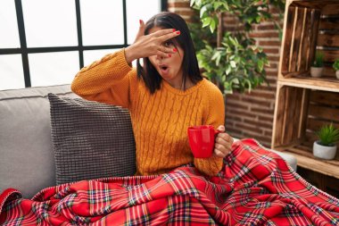 Young hispanic woman sitting on the sofa drinking a coffee at home peeking in shock covering face and eyes with hand, looking through fingers with embarrassed expression. 