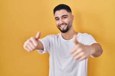 Sarı arka plan üzerine gündelik tişört giyen genç yakışıklı el hareketlerini olumlu karşılıyor, gülümsüyor ve başarı için mutlu. kazanan jest. 