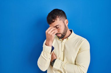 Handsome hispanic man standing over blue background tired rubbing nose and eyes feeling fatigue and headache. stress and frustration concept. 