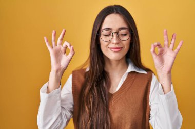 Sarı arka planda duran genç esmer kadın gözlük takıyor rahatlıyor ve gözleri kapalı gülümsüyor parmaklarıyla meditasyon hareketi yapıyor. yoga konsepti. 