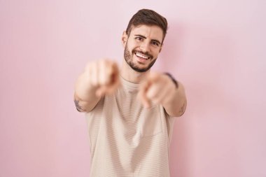 Sakallı İspanyol bir adam pembe arka planda seni ve kamerayı işaret ediyor, gülümsüyor, pozitif ve neşeli. 