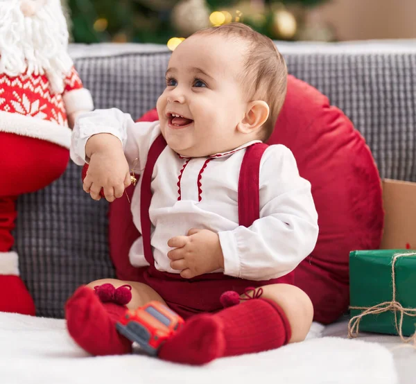 可爱的他的惊恐的宝宝自信地微笑着坐在家里的圣诞树旁的沙发上 — 图库照片