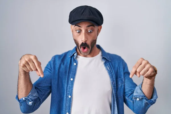 Hispanic man with beard standing over isolated background pointing down with fingers showing advertisement, surprised face and open mouth