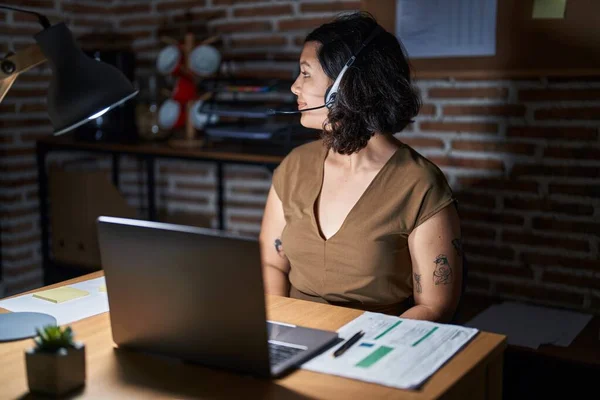 Young hispanic woman working at the office at night looking to side, relax profile pose with natural face and confident smile.