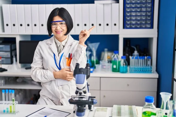 Young asian woman working at scientist laboratory smiling happy pointing with hand and finger to the side