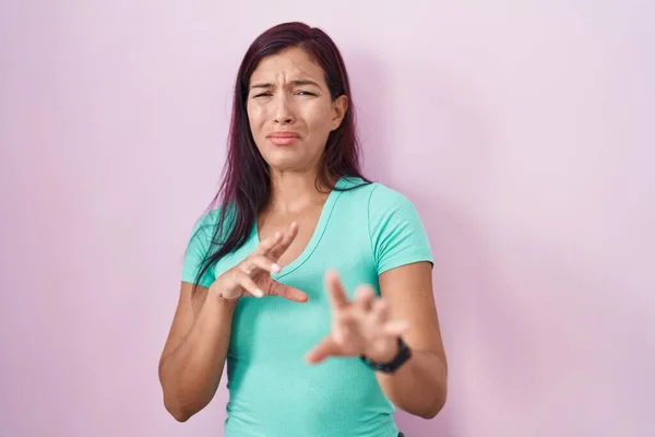 Young hispanic woman standing over pink background disgusted expression, displeased and fearful doing disgust face because aversion reaction.