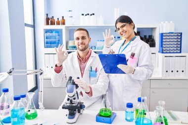 Laboratuvarda çalışan iki genç insan parmaklarıyla imza atıp gülümsüyor ve mükemmel bir sembol oluşturuyor. 