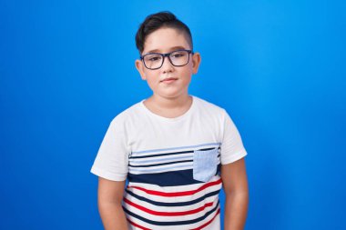 Young hispanic kid standing over blue background relaxed with serious expression on face. simple and natural looking at the camera. 