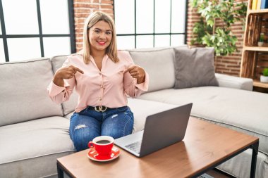 Young hispanic woman using laptop sitting on the sofa at home looking confident with smile on face, pointing oneself with fingers proud and happy. 