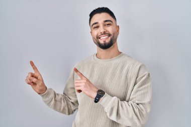 Genç yakışıklı adam izole edilmiş arka planda duruyor gülümsüyor ve kameraya bakıyor iki eliyle ve parmaklarıyla kenarı işaret ediyor.. 