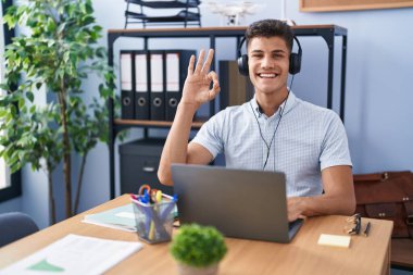 Young hispanic man working at the office wearing headphones smiling positive doing ok sign with hand and fingers. successful expression. 