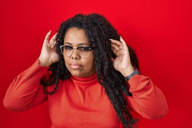 Plus size hispanic woman standing over red background trying to hear both hands on ear gesture, curious for gossip. hearing problem, deaf 