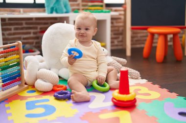 Anaokulunun zemininde duran sevimli beyaz bebek elinde çember oyuncağı.