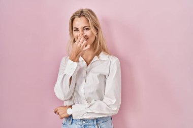 Young caucasian woman wearing casual white shirt over pink background smelling something stinky and disgusting, intolerable smell, holding breath with fingers on nose. bad smell 
