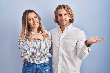Young couple standing over blue background amazed and smiling to the camera while presenting with hand and pointing with finger. 