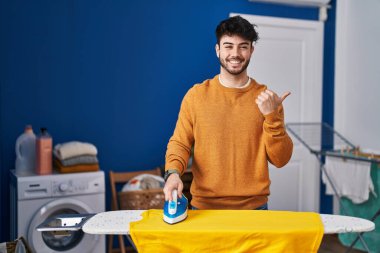 Evde giysilerini ütüleyen İspanyol sakallı bir adam baş parmağıyla ağzını açarak gülümsüyor. 