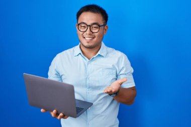 Chinese young man using computer laptop pointing aside with hands open palms showing copy space, presenting advertisement smiling excited happy 