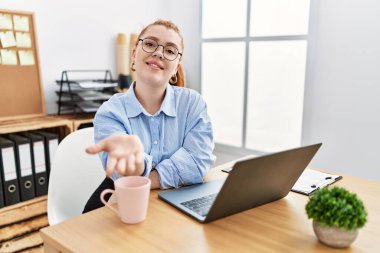 Young redhead woman working at the office using computer laptop smiling cheerful offering palm hand giving assistance and acceptance. 
