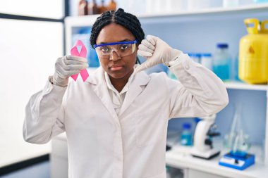 African american woman working at scientist laboratory holding pink ribbon with angry face, negative sign showing dislike with thumbs down, rejection concept 