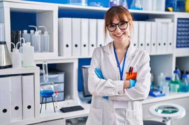 Genç bilim kadını gülümsüyor. Kollarını kavuşturup laboratuvarda duruyor.