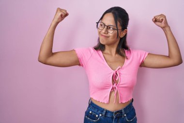 Hispanic young woman standing over pink background wearing glasses showing arms muscles smiling proud. fitness concept. 
