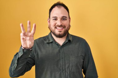Plus size hispanic man with beard standing over yellow background showing and pointing up with fingers number three while smiling confident and happy. 