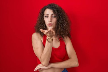 Hispanic woman with curly hair standing over red background looking at the camera blowing a kiss with hand on air being lovely and sexy. love expression. 