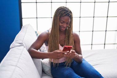 Evdeki koltukta oturan Afro-Amerikan kadın akıllı telefon kullanıyor