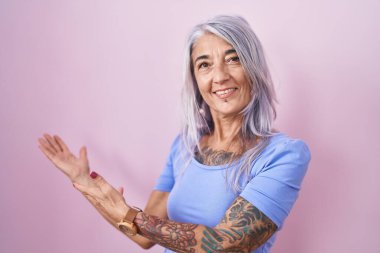 Pembe arka planda duran orta yaşlı bir kadın gülümseyerek doğal bir şekilde içeri girmeye davet ediyor. 