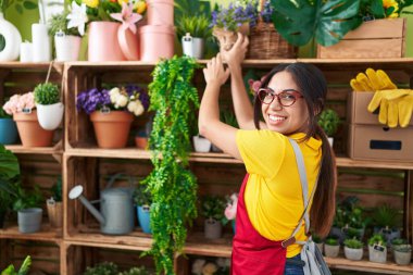 Young beautiful arab woman florist smiling confident holding plant of shelving at flower shop