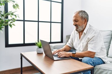 Evde dizüstü bilgisayar kullanarak gülümseyen yaşlı gri saçlı adam.