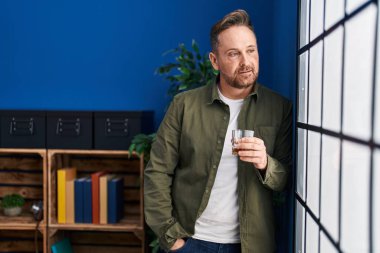 Genç beyaz adam evde dikilip bir bardak viski içiyor.