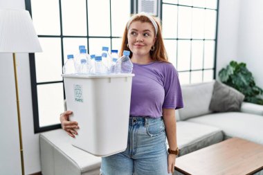 Young redhead woman holding recycling wastebasket with plastic bottles smiling looking to the side and staring away thinking. 