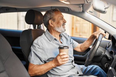 Yaşlı, kır saçlı, kahve içen adam caddede arabada oturuyor.