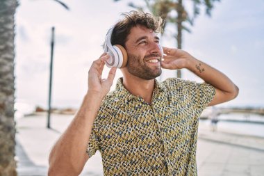 Genç İspanyol adam deniz kenarında müzik dinlerken kendinden emin bir şekilde gülümsüyor.