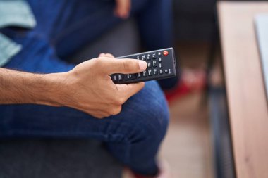 Young hispanic man holding tv remote control sitting on sofa at home