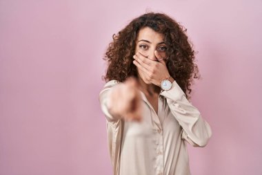 Hispanic woman with curly hair standing over pink background laughing at you, pointing finger to the camera with hand over mouth, shame expression 