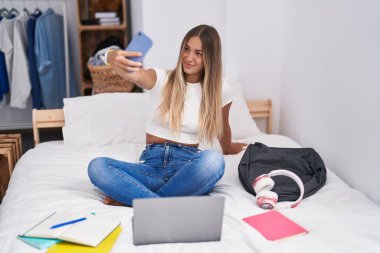 Young beautiful hispanic woman student make selfie by smartphone sitting on bed at bedroom