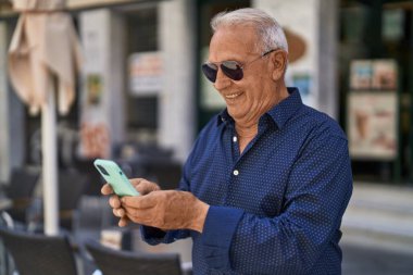 Yaşlı, gri saçlı, kendine güvenen, caddede akıllı telefon kullanan bir adam.