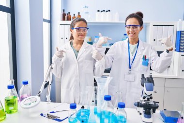 Young mother and daughter at scientist laboratory looking confident with smile on face, pointing oneself with fingers proud and happy. 