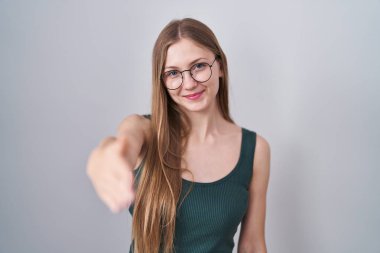 Young caucasian woman standing over white background smiling friendly offering handshake as greeting and welcoming. successful business. 