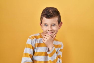 Young caucasian kid standing over yellow background looking confident at the camera smiling with crossed arms and hand raised on chin. thinking positive. 