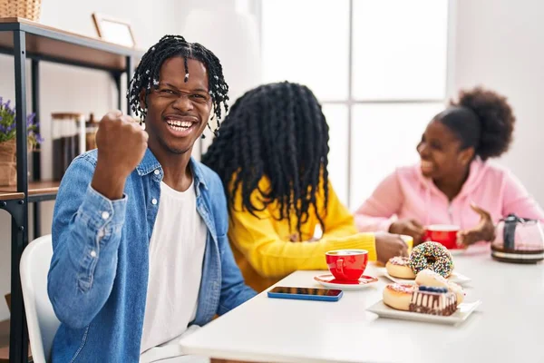 Group of three young black people sitting on a table having coffee screaming proud, celebrating victory and success very excited with raised arms