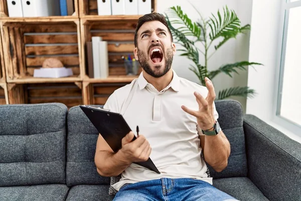 Handsome hispanic man holding clipboard working at psychology clinic crazy and mad shouting and yelling with aggressive expression and arms raised. frustration concept.