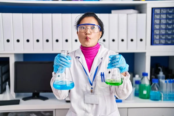 Young asian woman working at scientist laboratory making fish face with mouth and squinting eyes, crazy and comical.