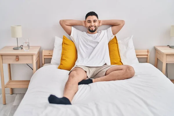Young arab man relaxed with hands on head sitting on bed at bedroom