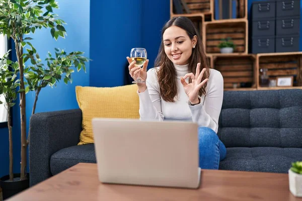 Young hispanic woman doing video call drinking white wine doing ok sign with fingers, smiling friendly gesturing excellent symbol