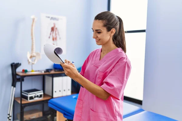 Young Hispanic Woman Wearing Physiotherapist Uniform Writing Clipboard Physiotherpy Clinic — Foto de Stock